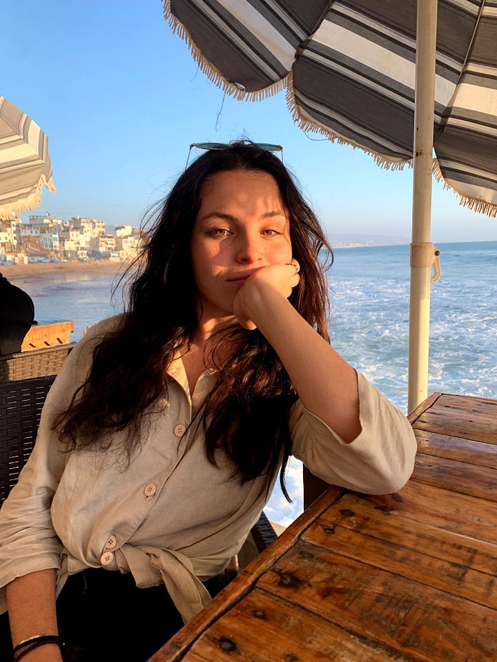 Split image: left, a woman with dark wavy hair resting her chin on her hand at a beachside café with the ocean and coastal buildings behind her; right, the same woman sitting on the wooden step of a white yurt under a partly cloudy sky.