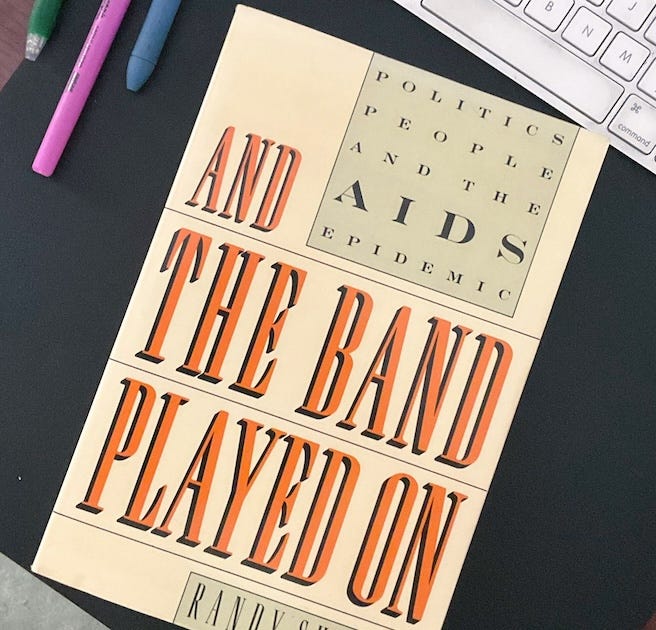 A copy of And the Band Played On: Politics, People, and the AIDS Epidemic by Randy Shilts lies on a dark tabletop. The book’s bold title, printed in orange and black lettering, dominates the cream-colored cover. A computer keyboard and three pens (pink, green, and purple) are partially visible in the frame, suggesting a workspace or moment of study.