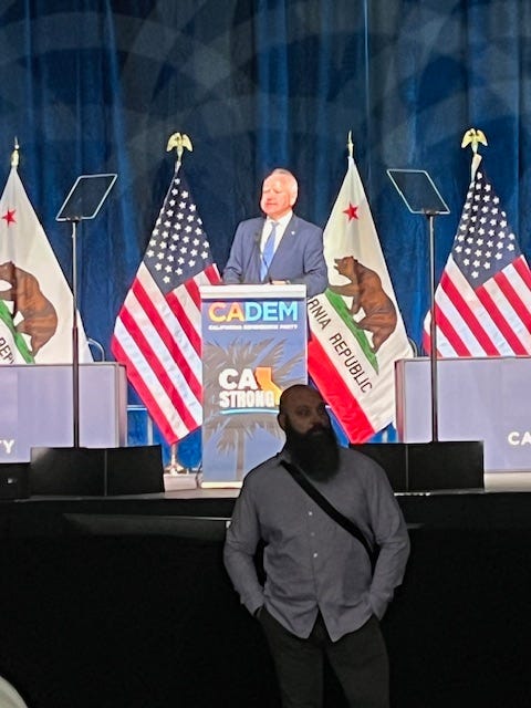 Gov. Tim Walz at a podium on stage in front of American flags. A bodyguard stands on the floor below in front of him.