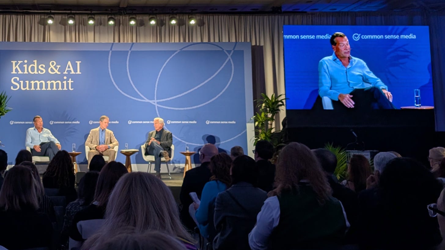 Three men seated on stage at the Kids & AI Summit, with one panelist shown on a large screen Three men seated on stage at the Kids & AI Summit, with one panelist shown on a large screen