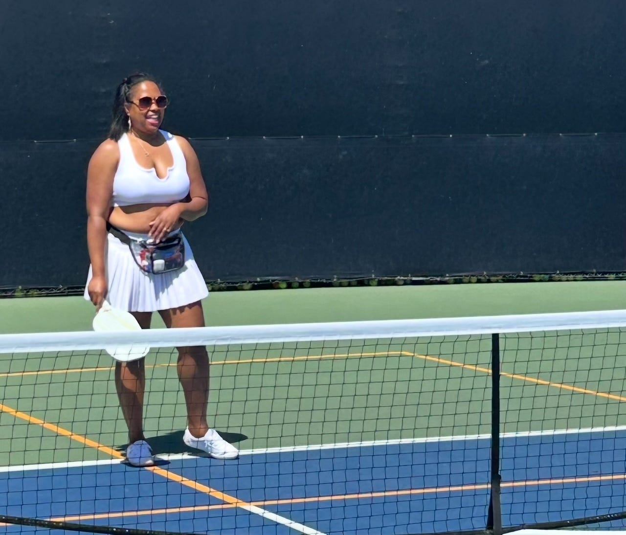 Black woman smiling in sunglasses and white top and skirt on pickleball court holding racket