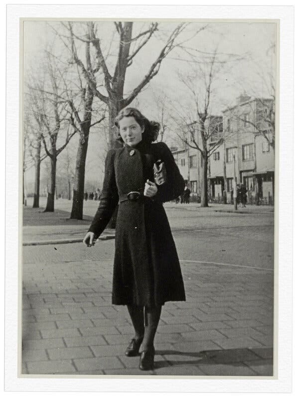 A black-and-white photo of Hannie Schaft wearing a long coat and standing in front of bare trees. A row of buildings is in the distance.