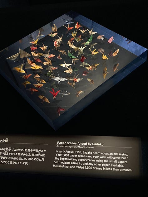 Photo of the Children's Peace Memorial, a tall concrete structure with a crane on top. Colorful strings of folded cranes. Museum display with tiny cranes foleded by Sasaki.