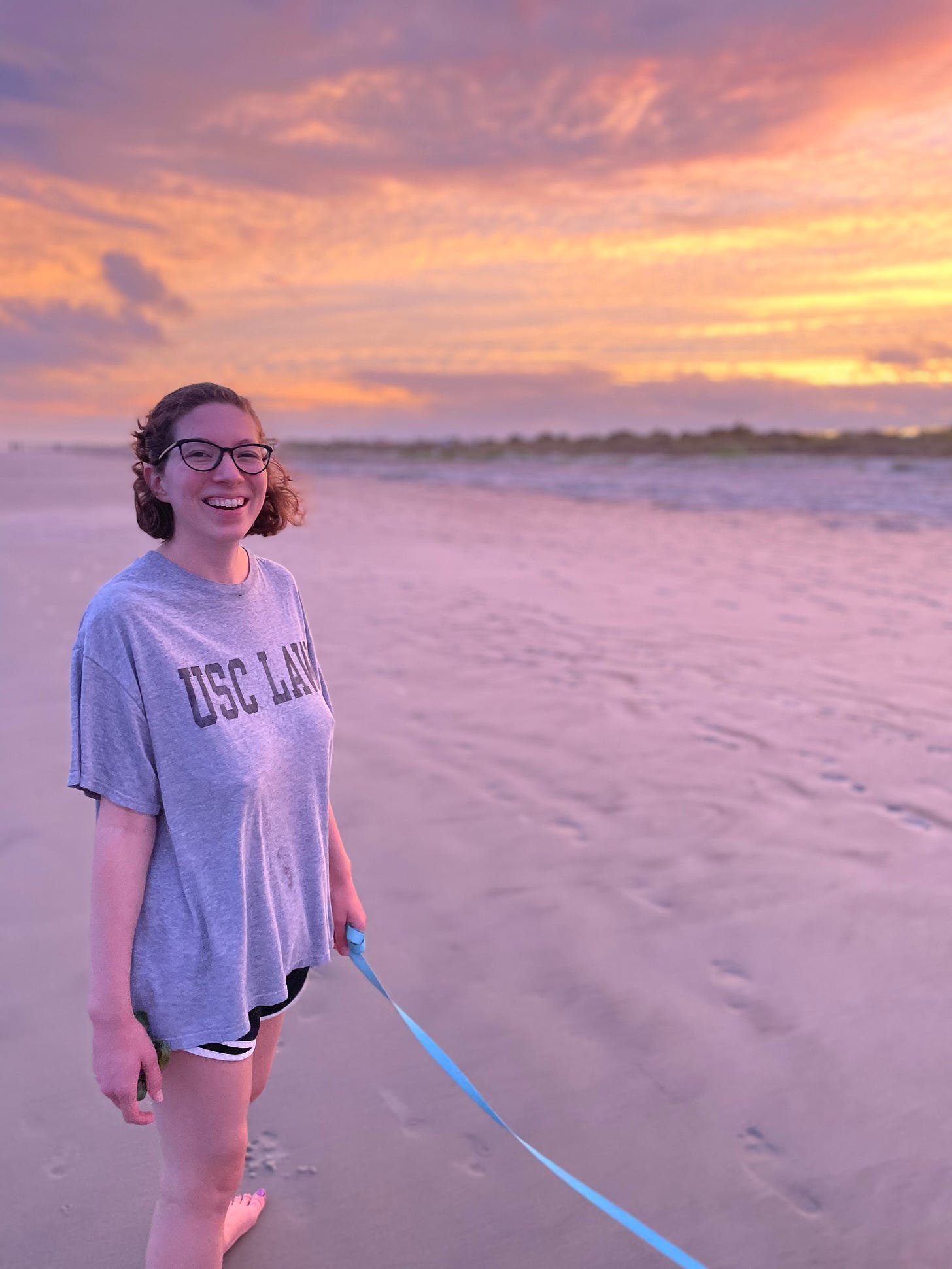 Image description: a white woman stands on the side of the frame and smiles at the camera. She is on a beach and the sun is setting. She has glasses and holds a dog leash, wearing a gray t-shirt that says "USC law" and black shorts.