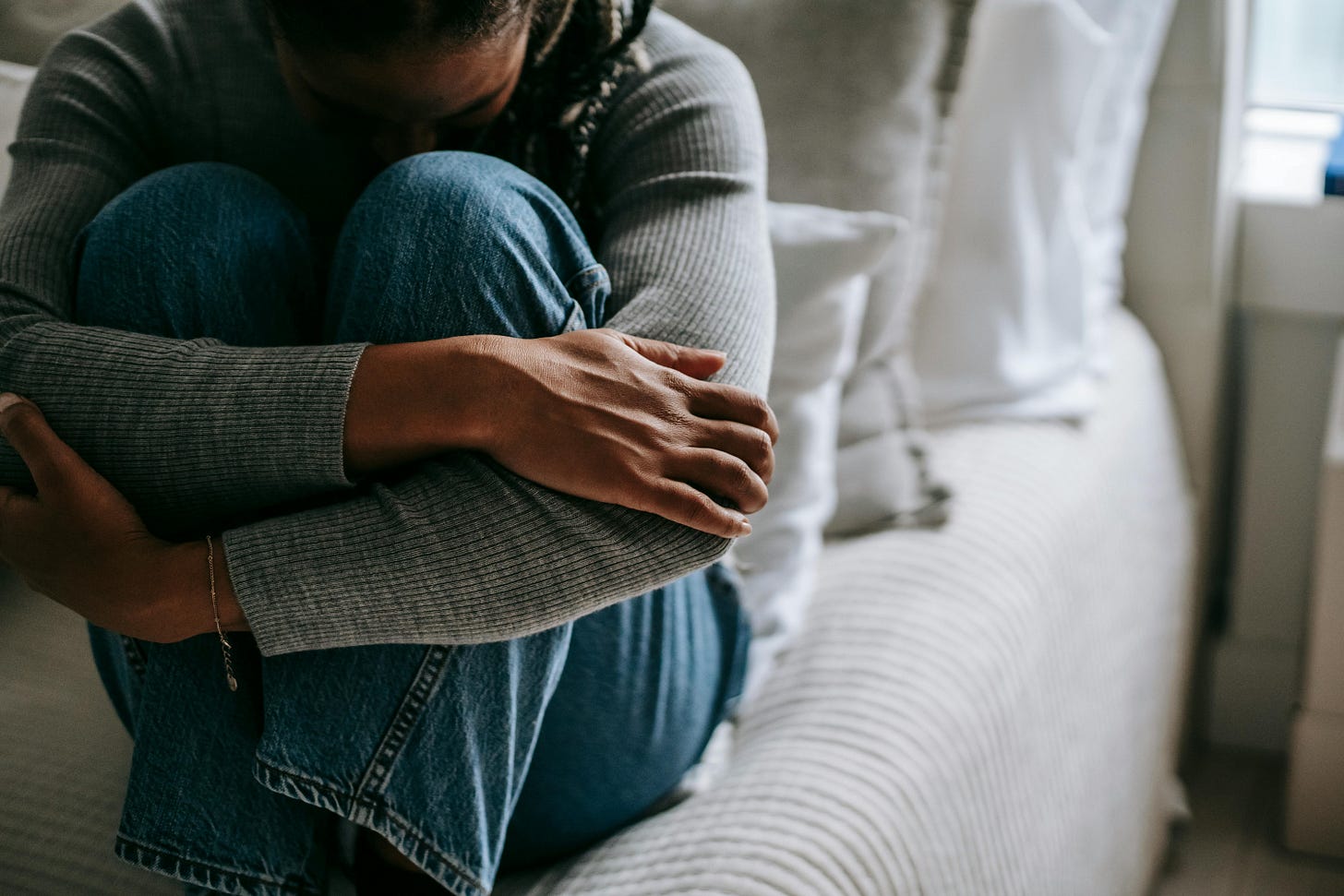 Close-Up von einer jungen Frau in Jeans und Pullover, die zusammengekauert auf einem Bett sitzt.