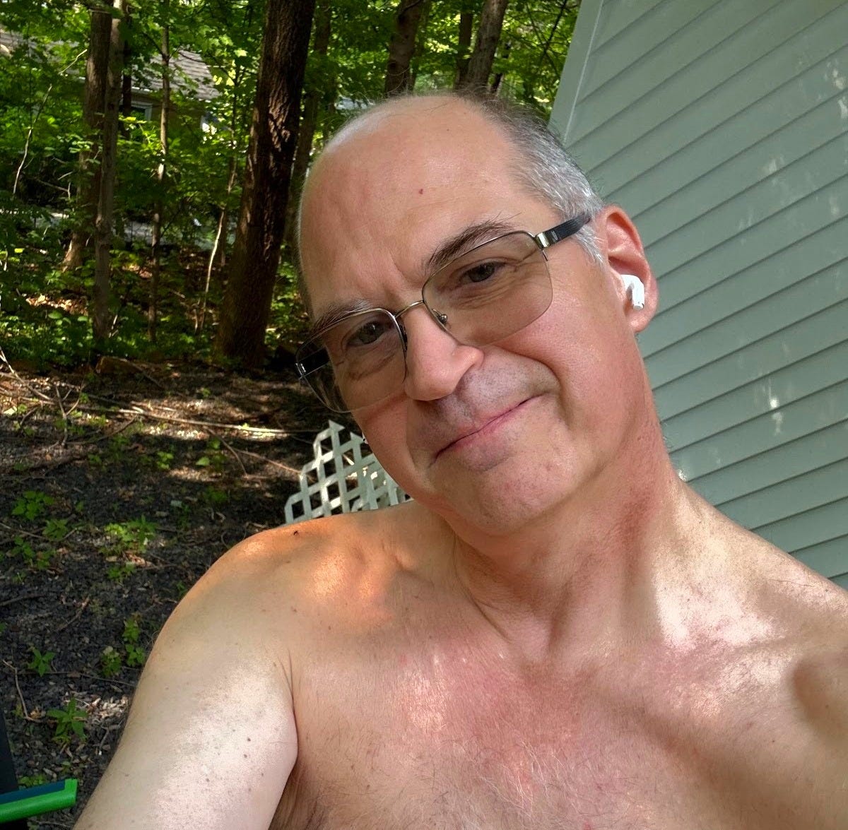 Dan Speers, known as Maker of Things, taking a topless selfie outdoors at Sky Farm, wearing glasses and wireless earbuds, with trees and a building visible in the background.