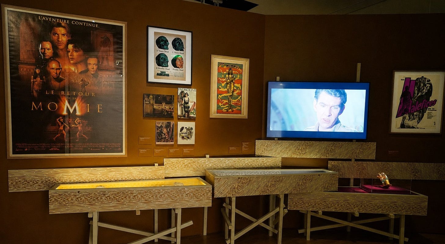 A Paris exhibition display on mummies in popular culture, including a poster for The Mummy, a video screen, and printed materials.