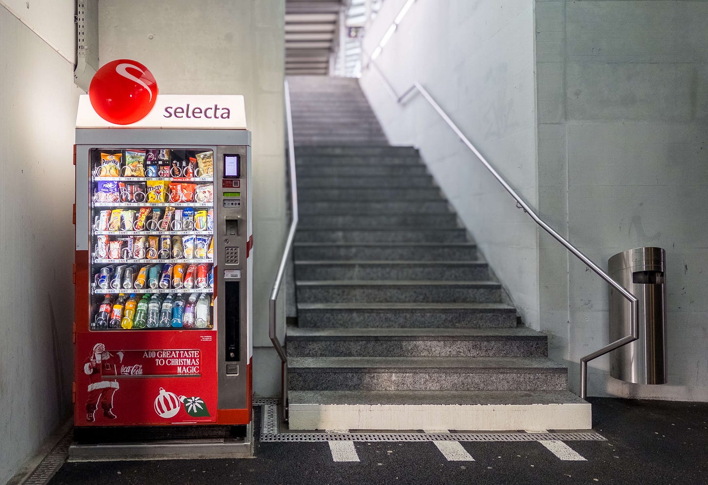 Das Foto zeigt einen Verkaufsautomaten der Marke Selecta, der Snacks und Getränke anbietet. Der Automat steht neben einer breiten Treppe in einer nüchternen, funktionalen Umgebung, wie sie typisch für Bahnhöfe oder öffentliche Plätze ist. Im oberen Bereich des Automaten prangt ein rotes Logo mit der Aufschrift „Selecta“. Der Automat ist gut gefüllt mit Chips, Schokoriegeln, Müsliriegeln, Softdrinks und Wasser. Unten auf dem Automaten befindet sich ein rotes Werbeplakat von Coca-Cola mit einem Weihnachtsmann und der Aufschrift „Add great taste to Christmas magic“. Rechts neben dem Automaten steht ein moderner Edelstahl-Mülleimer. Die Treppe im Hintergrund führt nach oben in einen helleren Bereich.