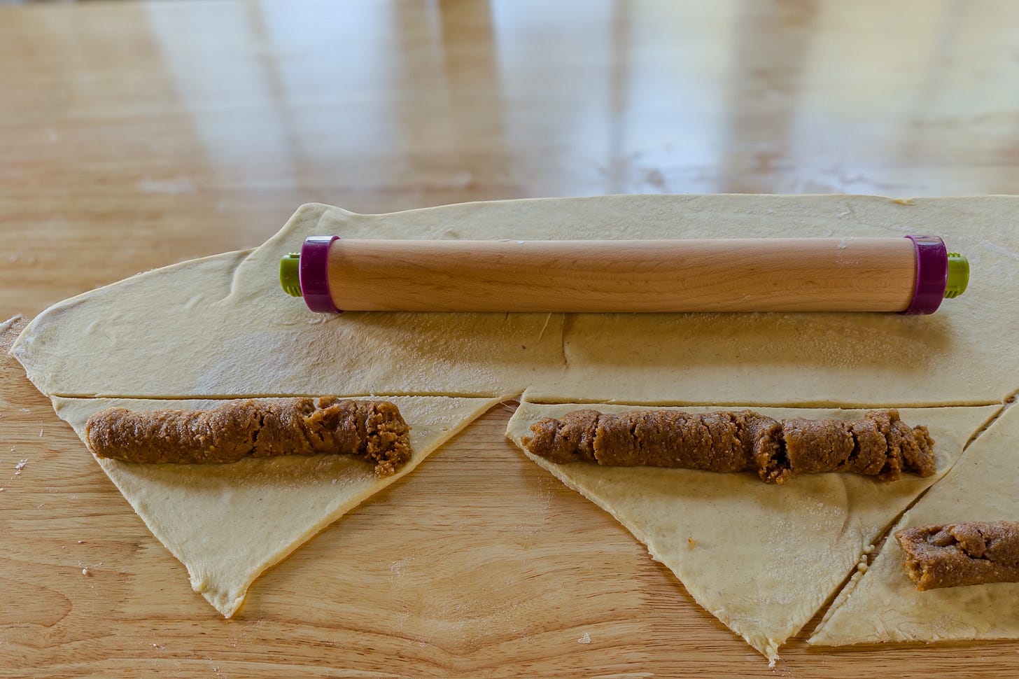 Ausgewallter Hefeteig für Nussgipfel auf einer Holzfläche, in Dreiecke geschnitten und mit länglichen Portionen Nussfüllung belegt. Ein Nudelholz liegt auf dem restlichen Teig. Ausgewallter Hefeteig für Nussgipfel auf einer Holzfläche, in Dreiecke geschnitten und mit länglichen Portionen Nussfüllung belegt. Ein Nudelholz liegt auf dem restlichen Teig.