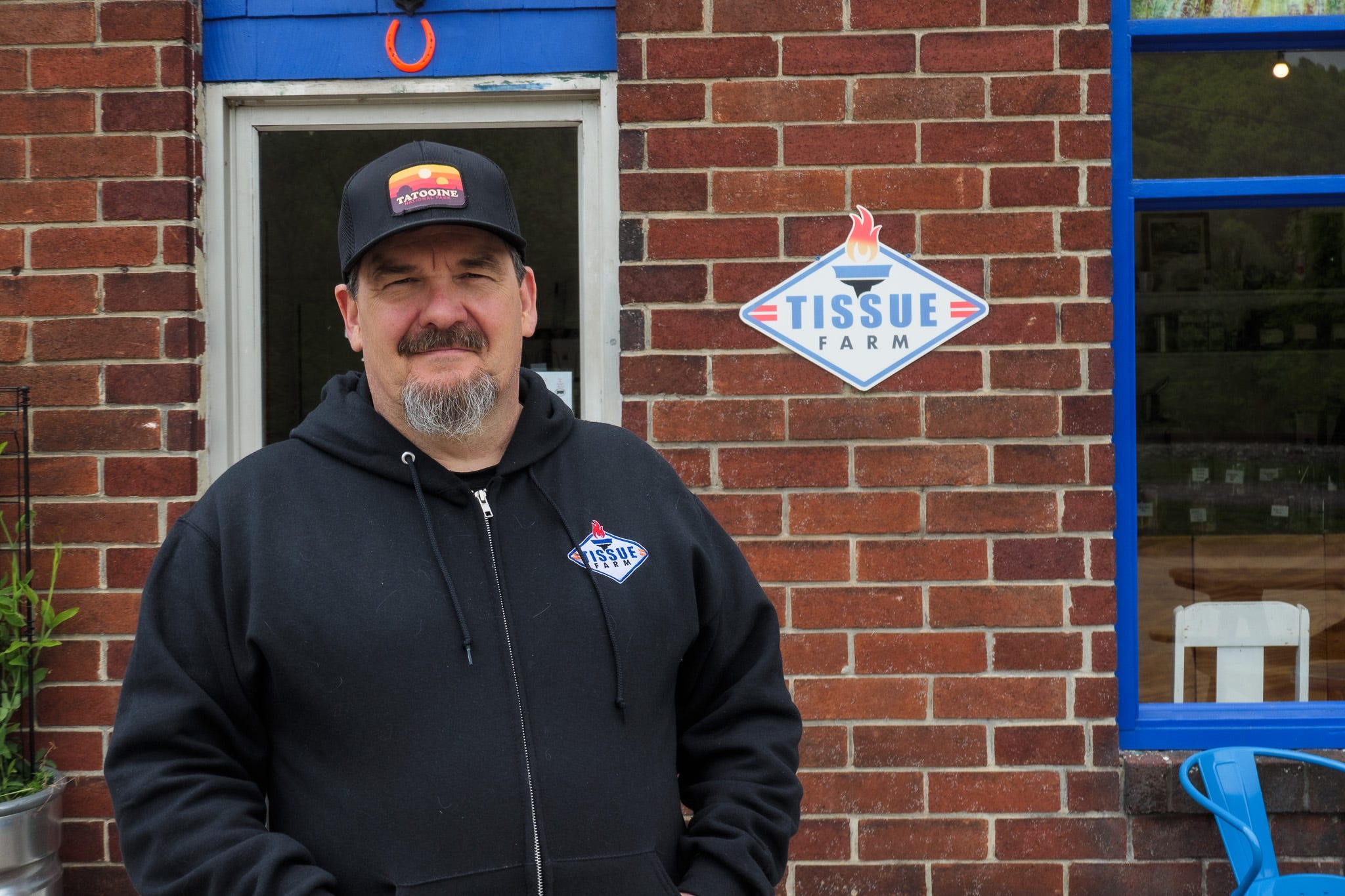 Photo of Pope, owner of Tissue Farm cafe and art gallery. Pope is middle-aged with a dark mustache and greying beard. Pope is wearing a black hoodie with the Tissue Farm logo - a flattened white diamond with a flaming torch and "TISSUE FARM" lettering. He is wearing a black baseball cap with a patch that features a desert landscape of yellow, orange, and brown, with the lettering "Tatooine". Behind him is the brick exterior wall of the cafe with the "Tissue Farm" logo on the wall.