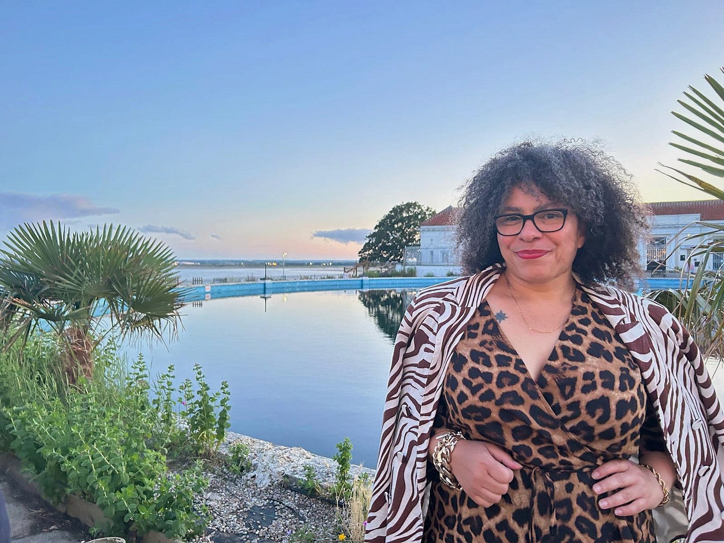 Susie in an animal print jacket and dress standing infront of a boating pool and the sea at sunset Susie in an animal print jacket and dress standing infront of a boating pool and the sea at sunset