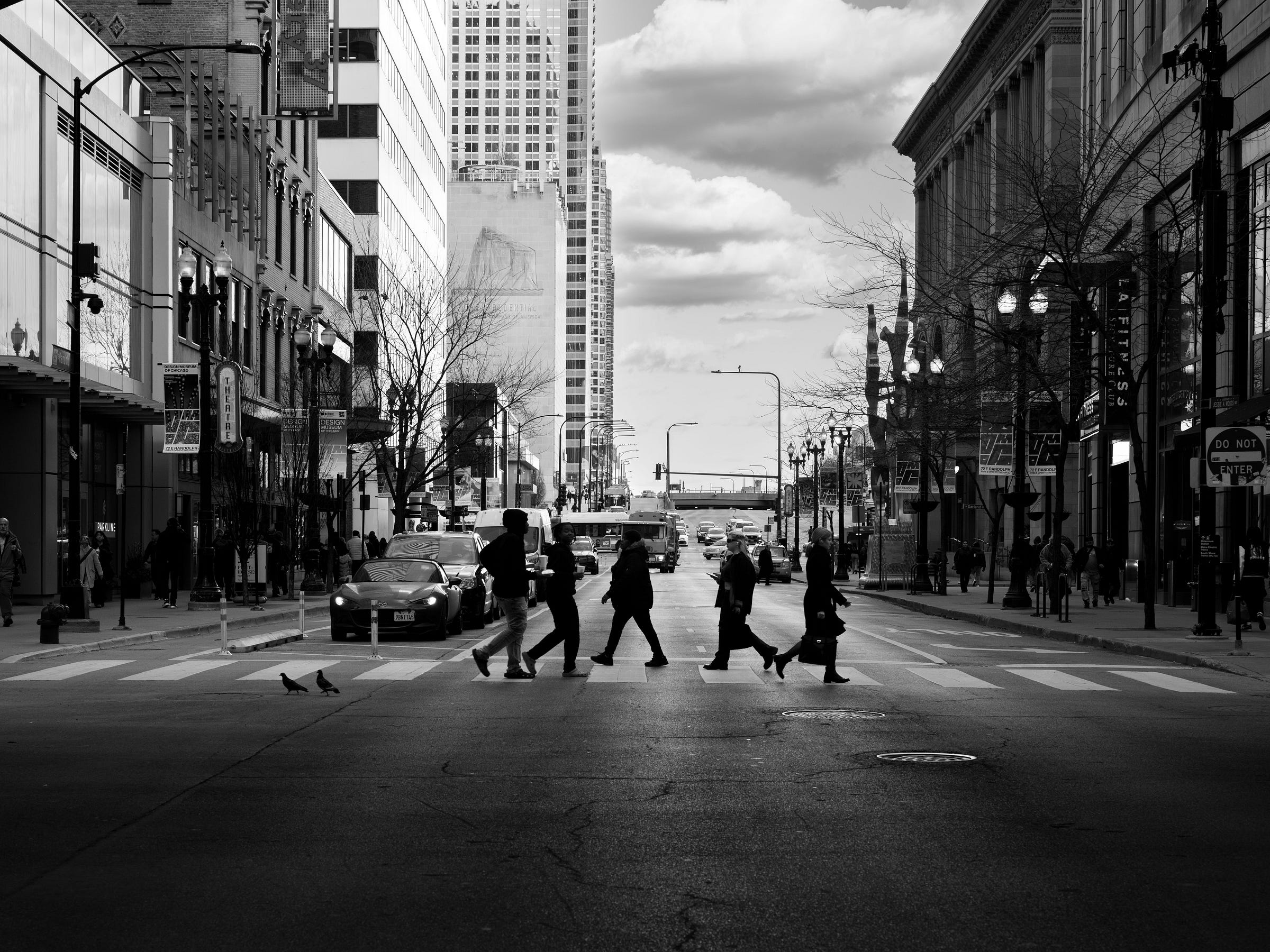 Black and white photograph of a busy city street with pedestrians crossing a crosswalk in silhouette. Cars wait at the intersection, and pigeons peck at the pavement in the foreground. The towering buildings, streetlights, and a cloudy sky frame the bustling urban scene. Black and white photograph of a busy city street with pedestrians crossing a crosswalk in silhouette. Cars wait at the intersection, and pigeons peck at the pavement in the foreground. The towering buildings, streetlights, and a cloudy sky frame the bustling urban scene.