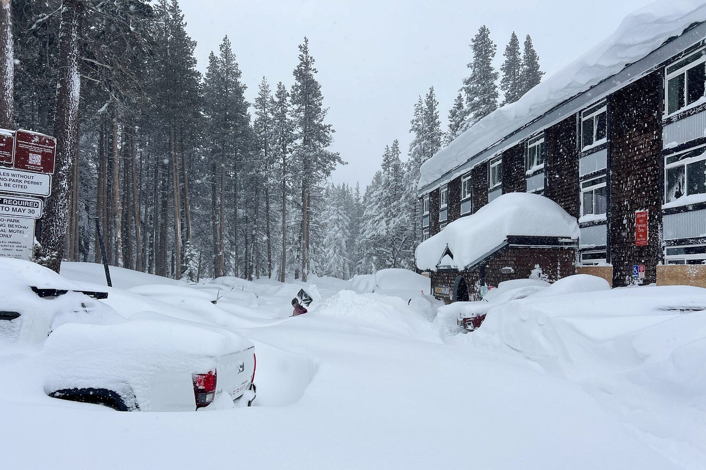 A snow-covered parking lot and motel building during heavy snowfall in a forested mountain area, with vehicles and structures buried under several feet of snow.