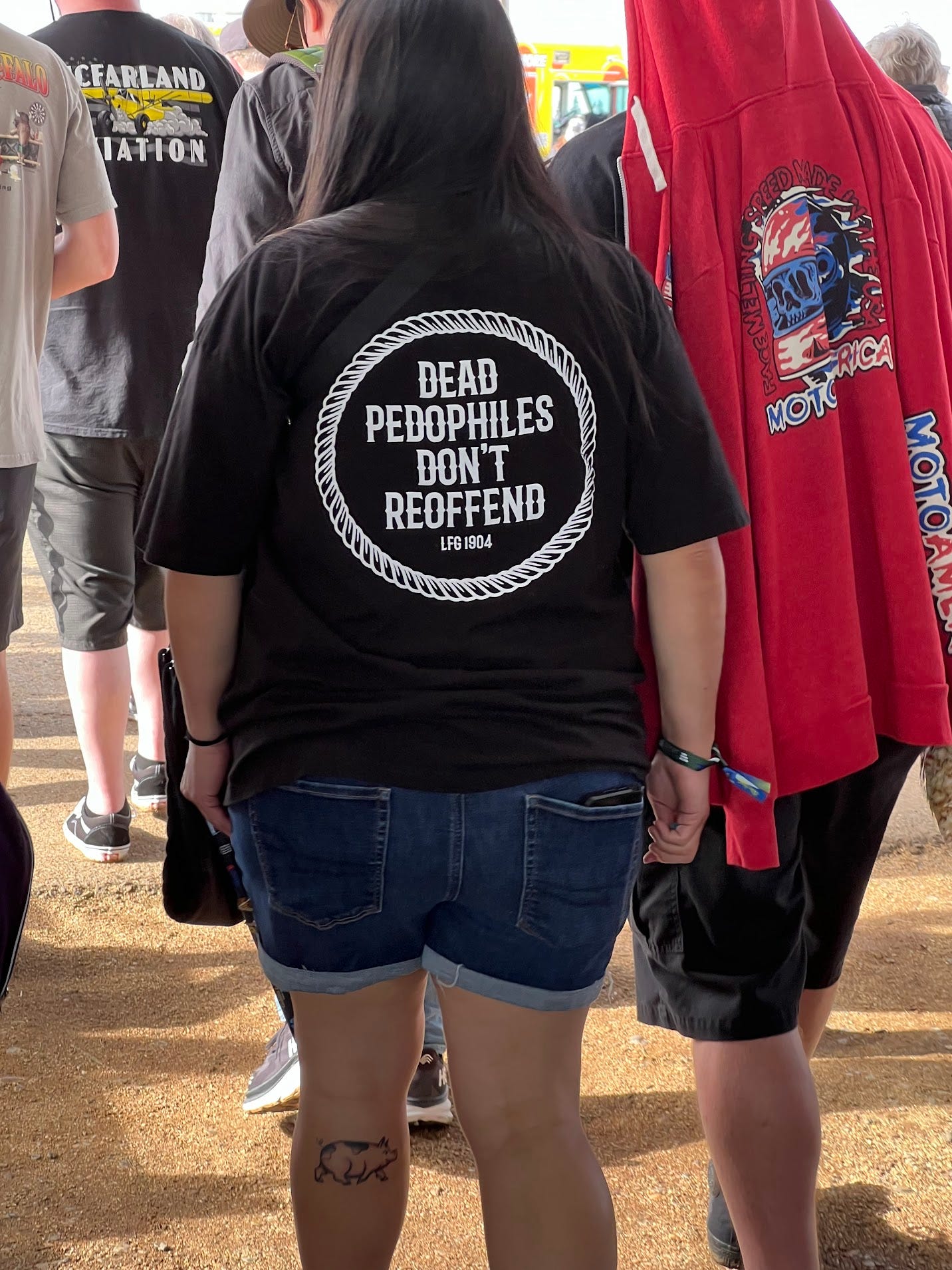 A photograph of a young woman from behind. She is wearing a black T-shirt with an image composed of a ring in which is the slogan, “Dead Pedophiles Don’t Reoffend,” and the inscription, “LFG1904.” She has a tattoo of a cartoon pig at the top of her left calf. Her boyfriend, to her right, has a red hoodie draped from his head and down his back. The hoodie has a colorful logo of a skull in a flaming motorcycle helmet and the brand “Motoamerica” written in blue and red.