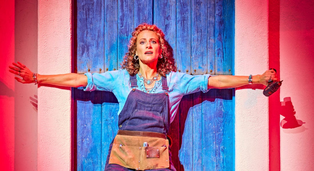 A woman wearing a blue shirt and dungarees with her arms outstretched by a door A woman wearing a blue shirt and dungarees with her arms outstretched by a door