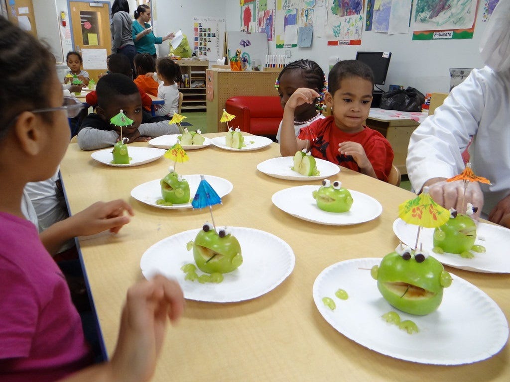 Children at a classroom table waiting to eat (or play with?) apple halves that have been decorated to resemble frogs, with sliced-grape 'feet.' The 'frogs' have googly eyes made with marshmallows and olive halves (yum!) attached to the apples with toothpicks. Children at a classroom table waiting to eat (or play with?) apple halves that have been decorated to resemble frogs, with sliced-grape 'feet.' The 'frogs' have googly eyes made with marshmallows and olive halves (yum!) attached to the apples with toothpicks.