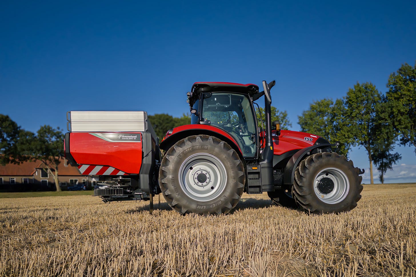 Ein roter Case IH-Traktor mit angebautem Kverneland Exacta-CL Düngerstreuer steht auf einem abgeernteten Feld. Im Hintergrund sind Bäume und Bauernhäuser zu sehen, darüber ein klarer blauer Himmel.