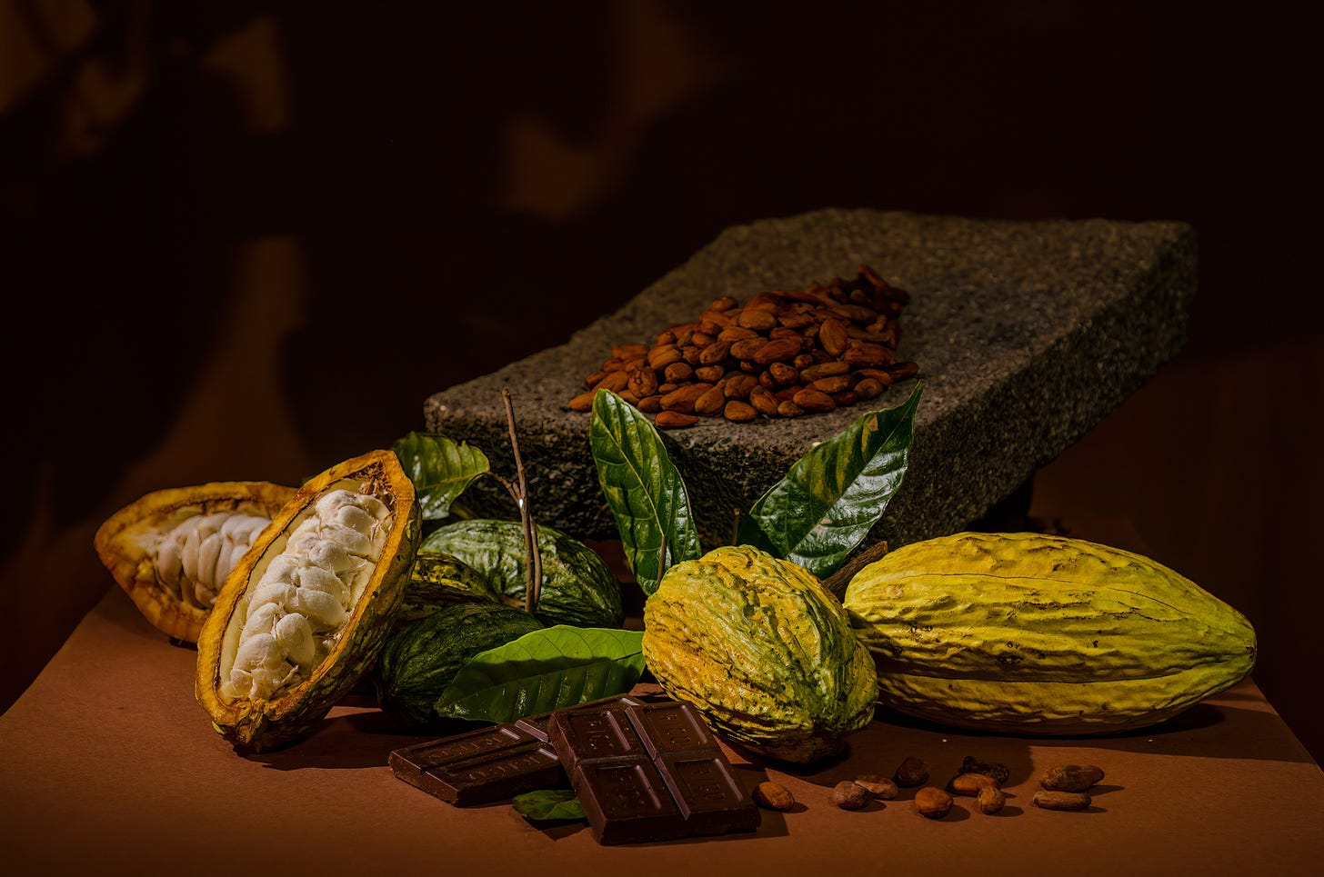 Assortment of cocoa-related items on a surface, including two whole cocoa pods—one cut open to reveal white pulp and seeds—scattered cocoa beans, a stone grinding slab, green leaves, and pieces of dark chocolate in the foreground, all set against a dark brown background