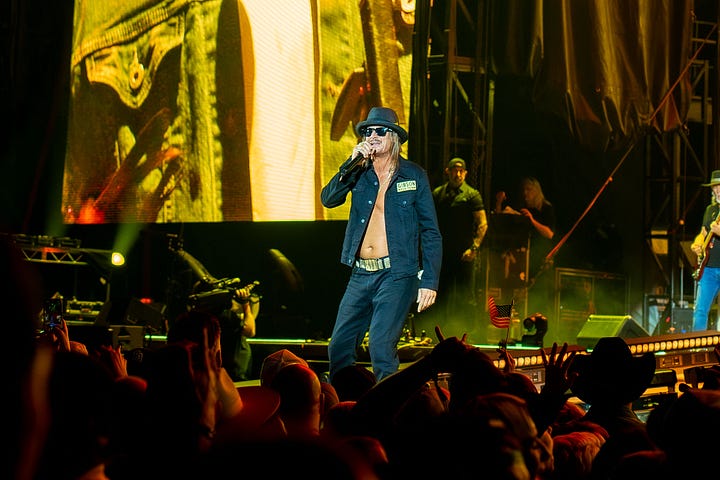 Kid rock is wearing a black open face button down, black hat and black sun glasses singing on stage with his band while the crowd dances to his music