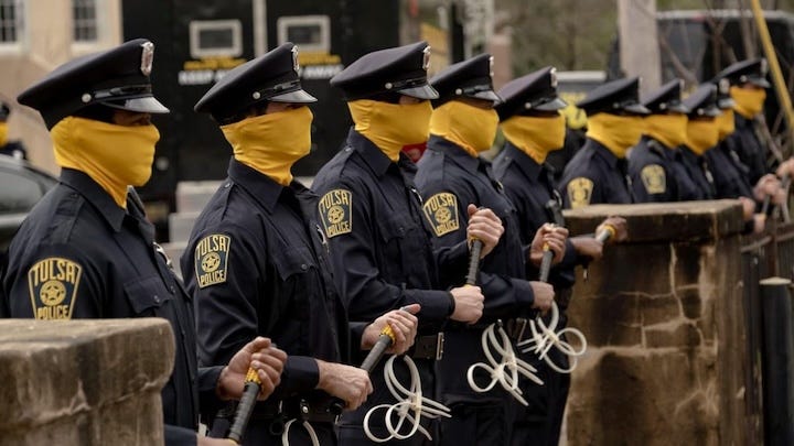 A lineup of Tulsa Police officers wearing yellow face coverings and dark uniforms, each holding zip-tie restraints. The visual evokes militarized order and masked authority—a central theme of the series.