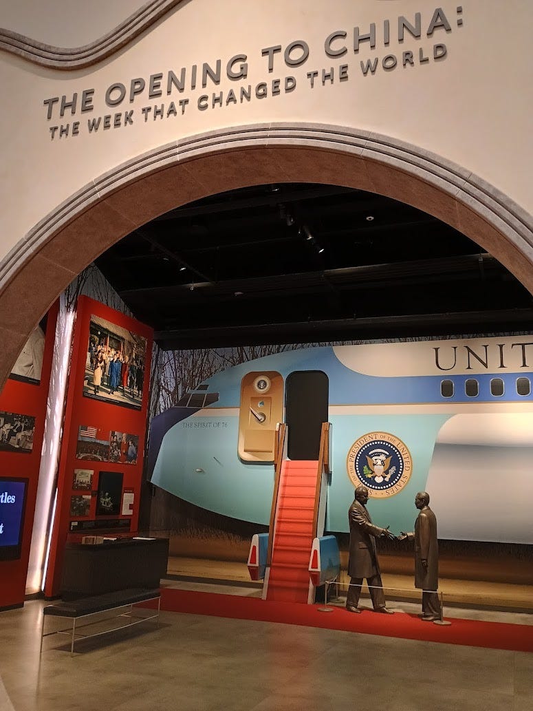 Exhibit titled The Opening to China: The Week That Changed the World, with giant image of Air Force One plane, and silhouette of Nixon extending hand to Chou Enlai. Pictures from China visit around walls.