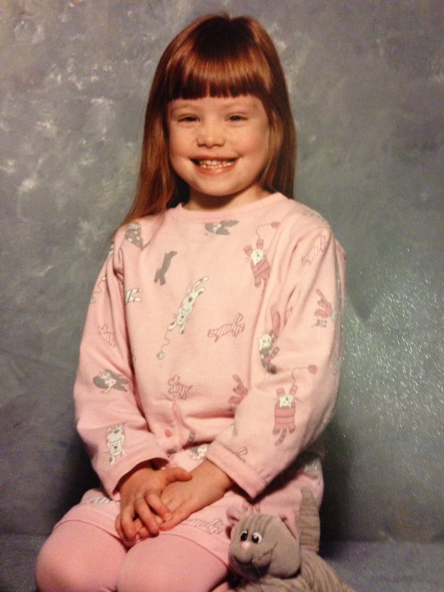A red-haired white kid wearing a pink sweatshirt dress with a playful cat pattern kneels in pink tights, hands in lap, with a stuffed gray cat by her side. 