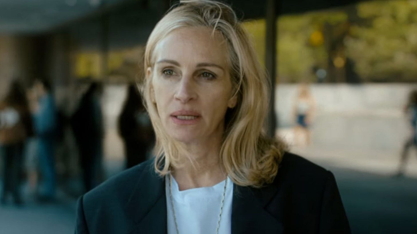 Mulher com cabelo loiro e longo, usando um blazer escuro, com um olhar sério em um ambiente externo. Ao fundo, várias pessoas são visíveis, mas desfocadas.