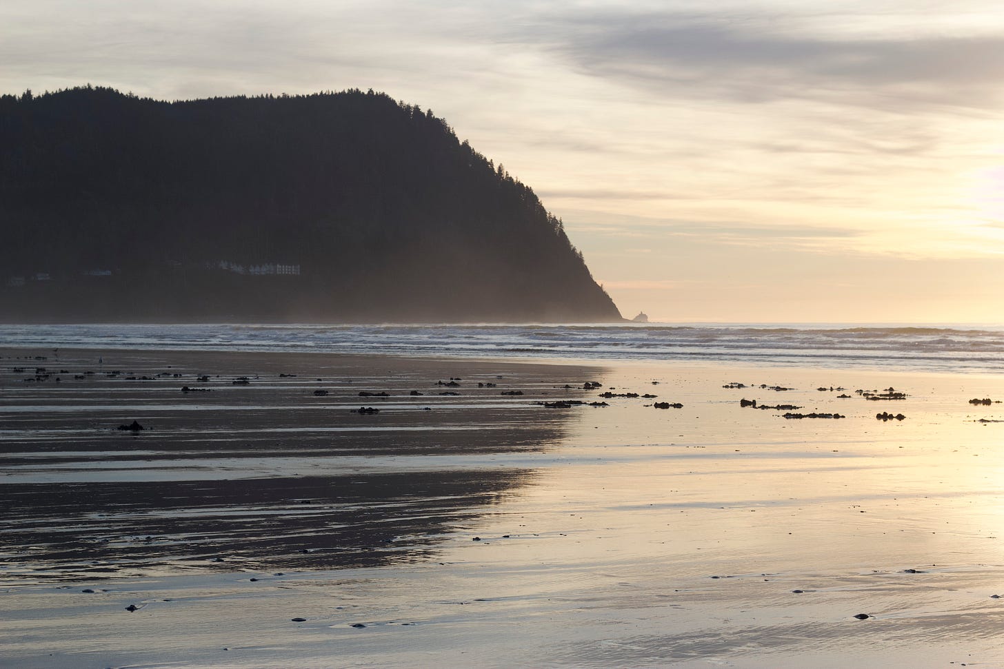 A partially cloudy sky just before sunset on a temperate late January day in 2022. Wet sand at low tide reflects the sun and the dark green of a steep hillside on the horizon looking southward from the beach at Seaside, Oregon.