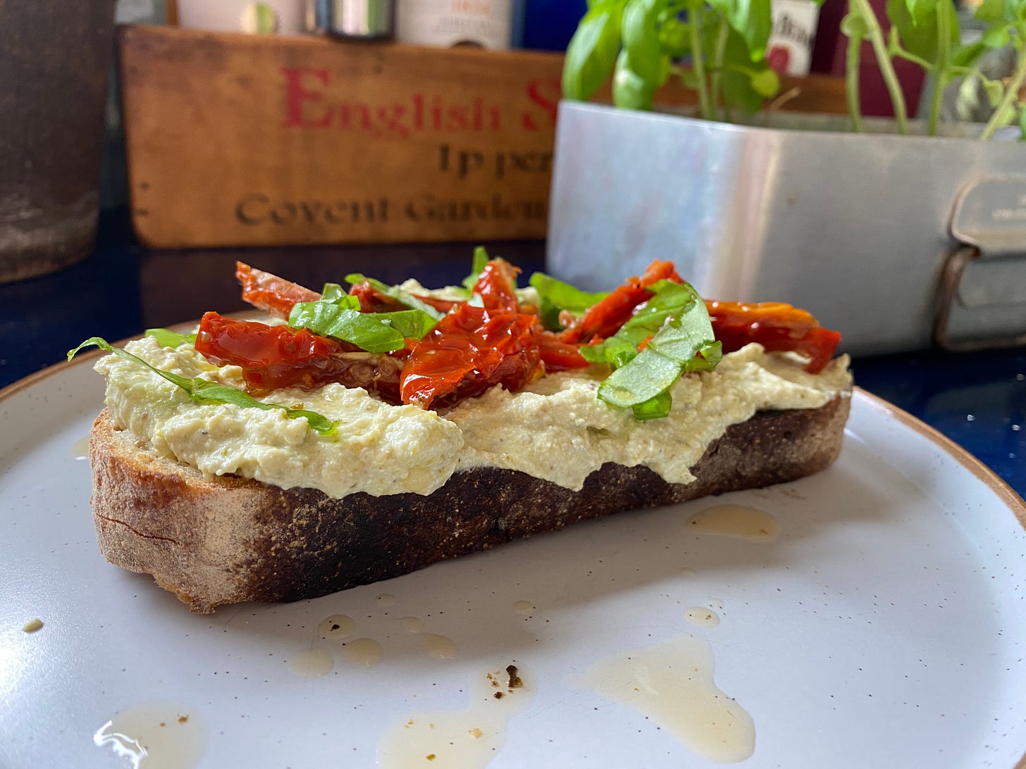 Slice of toast topped with cream cheese and sliced red sun-dried tomatoes and torn basil leaves. A silver container with basil plants is placed behind the plate on the worktop.