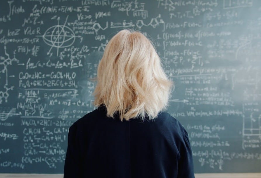 Woman with blonde hair facing a chalkboard with equations.