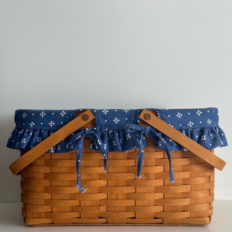 May include: A wooden picnic basket with blue and white fabric lining. The basket has two wooden handles and a woven pattern.