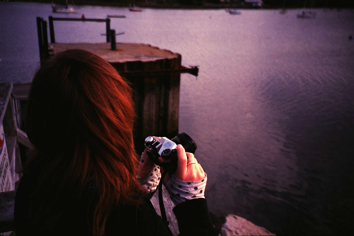A photograph of Sarah, with red hair, turned entirely away from the camera. Her arms seem to lean against a wooden beam just next to a body of water. In her hands, she holds a film camera. Polka-dotted sleeves peek out beneath her black jacket.