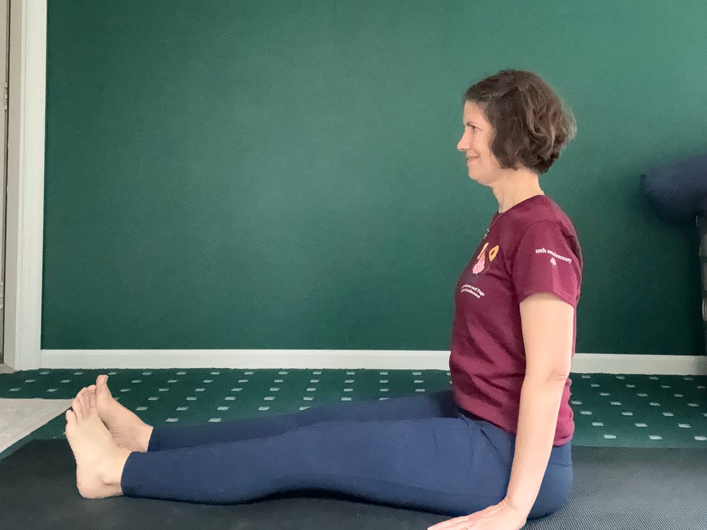 A middle-age woman with brown hair (me) sits upright on a black mat, legs stretched straight forward, arms at her side.
