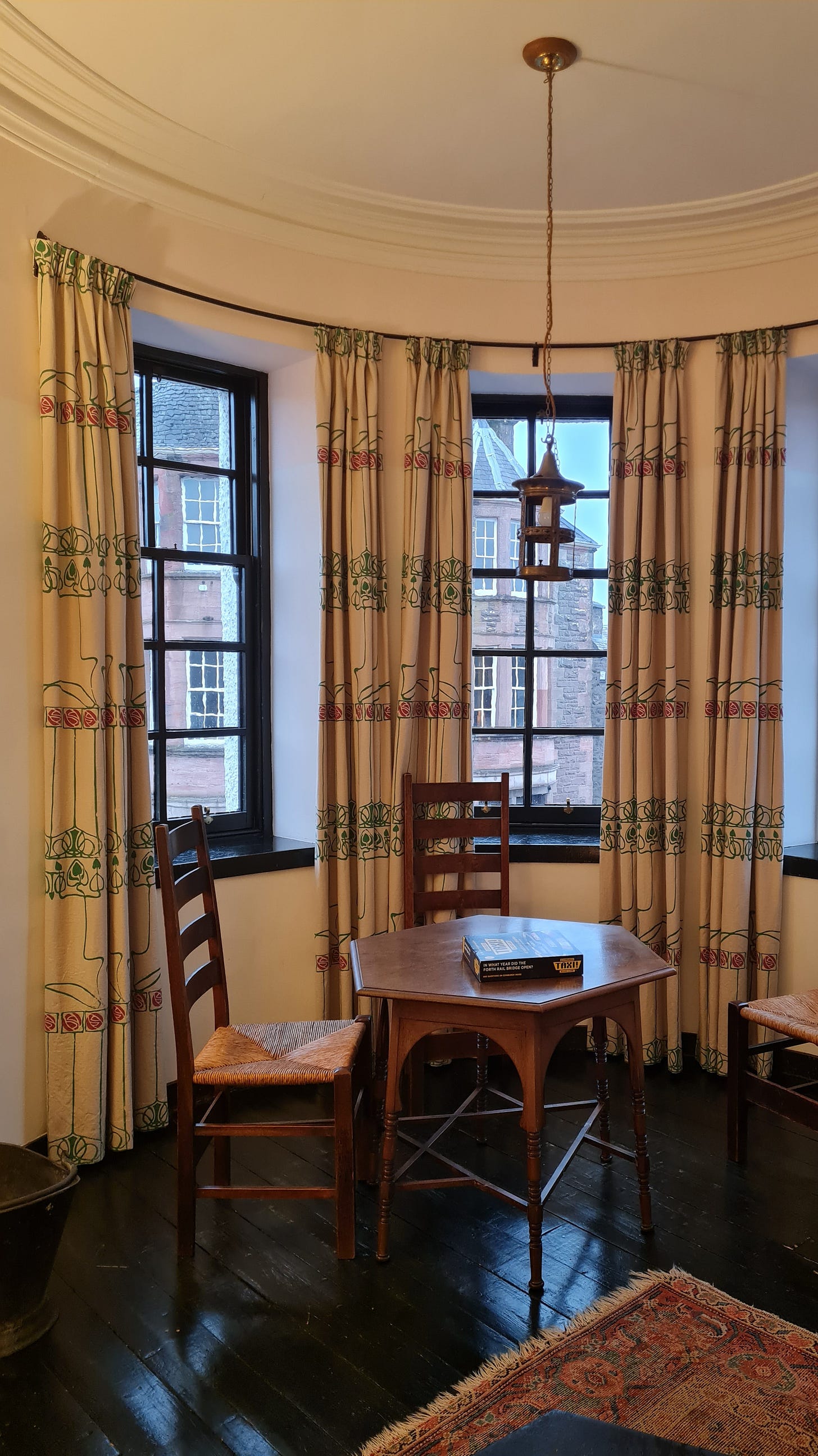 A room interior, with three windows with black wooden window frames. The walls are white and the floorboards are black. There's a small wooden table and three chairs with tall backs positioned round the table.