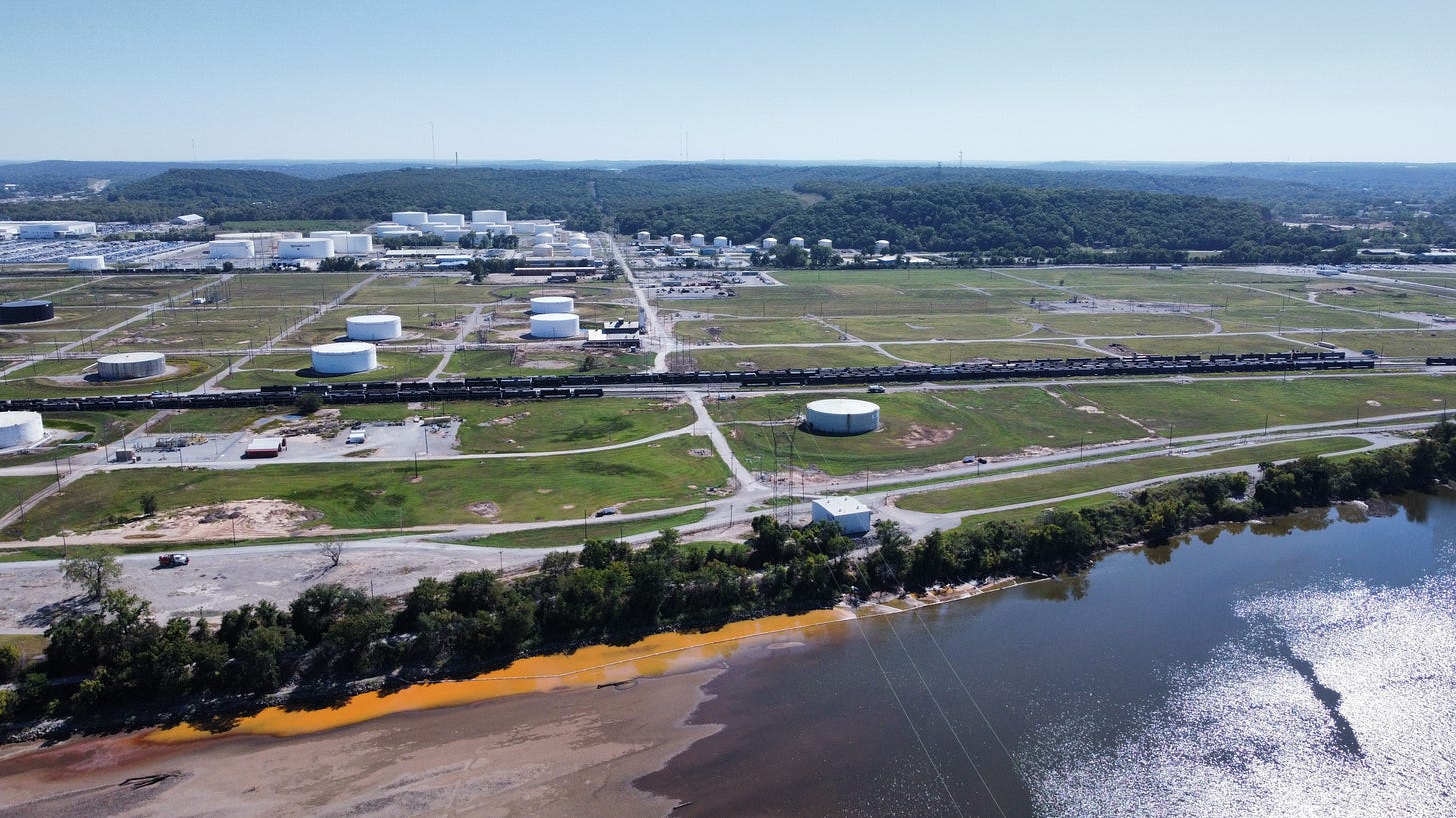 An opaque, bright orange plume escapes the bounds of a narrow stretch of white booms (tube-shaped floating barriers) along the Arkansas River shoreline. On the bank, a band of leafy trees casts shadows over the plume, which encroaches on a large sandbar and mixes with the dark green river water. In the background, an expanse of tank farms and grass lots cover the floodplain to the forested hills beyond.