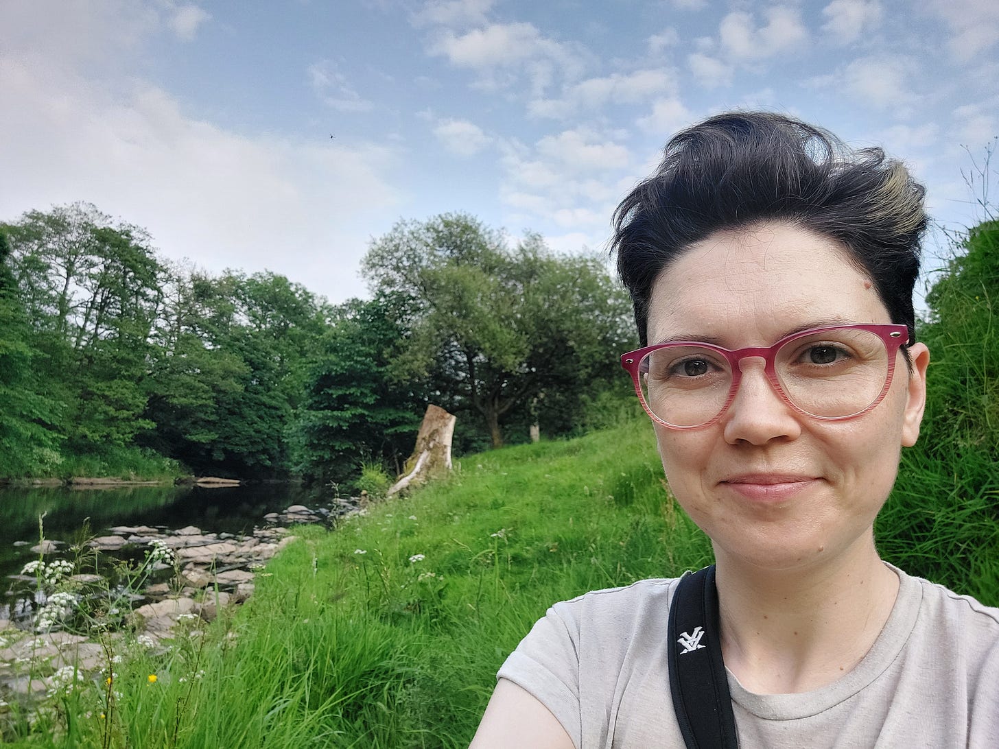 Selfie of Janelle (white woman with pixie short hair and pink glasses) sitting on some grass next to a rocky river