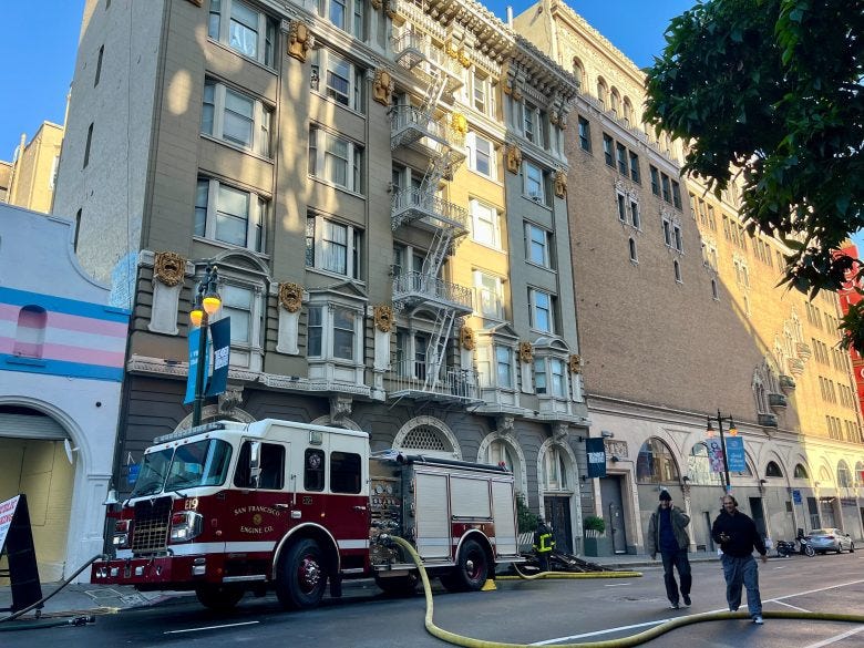 A San Francisco fire truck is parked outside a multi-story building with a hose extended, while two people walk by on the sidewalk. A San Francisco fire truck is parked outside a multi-story building with a hose extended, while two people walk by on the sidewalk.