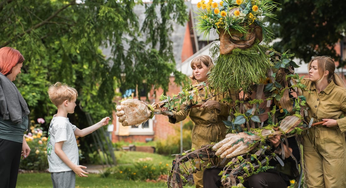 A large wooden puppet being managed by two women wearing boiler suits, reaching for the hand of a boy on the left