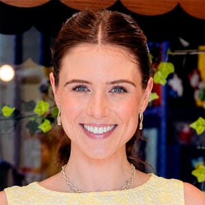 Mulher sorridente, com cabelos castanhos presos, olhos verdes, usando brincos, colar e blusa amarela, em frente a uma vitrine decorada Mulher sorridente, com cabelos castanhos presos, olhos verdes, usando brincos, colar e blusa amarela, em frente a uma vitrine decorada
