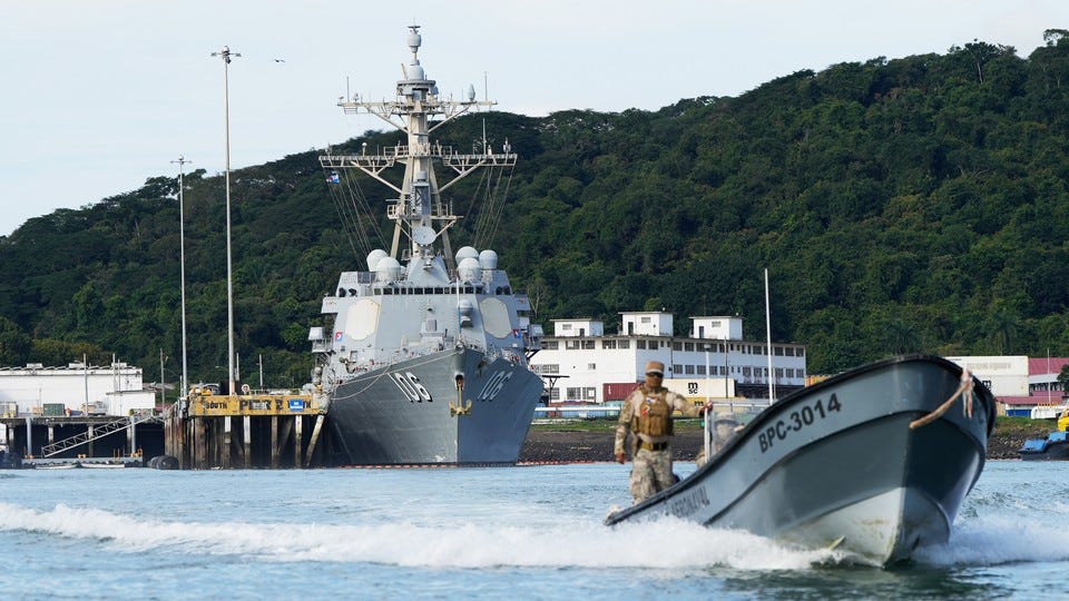 A man in camouflage drives a gray skiff away from a dock where a large, gray U.S. Navy ship sits.