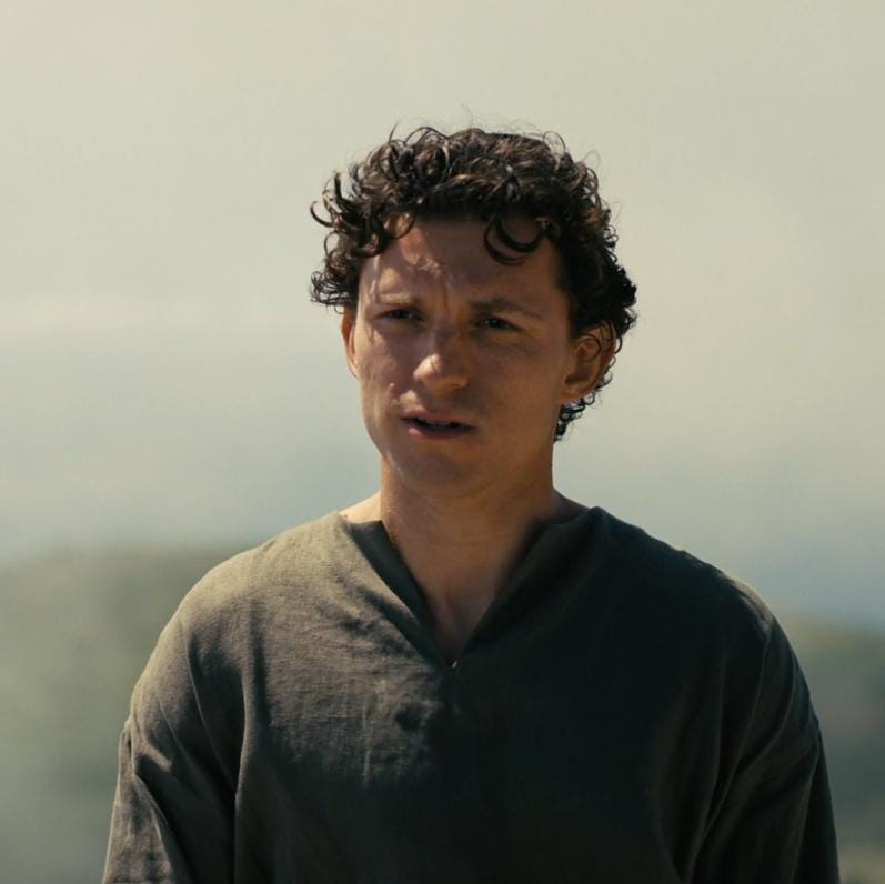 Telemachus, played by Tom Holland, stands outdoors in a plain gray tunic, looking thoughtful and uncertain. Telemachus, played by Tom Holland, stands outdoors in a plain gray tunic, looking thoughtful and uncertain.