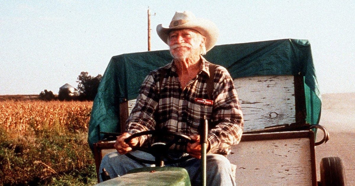 Elderly guy in a hat and button down shirt riding on a tractor beside farmland Elderly guy in a hat and button down shirt riding on a tractor beside farmland