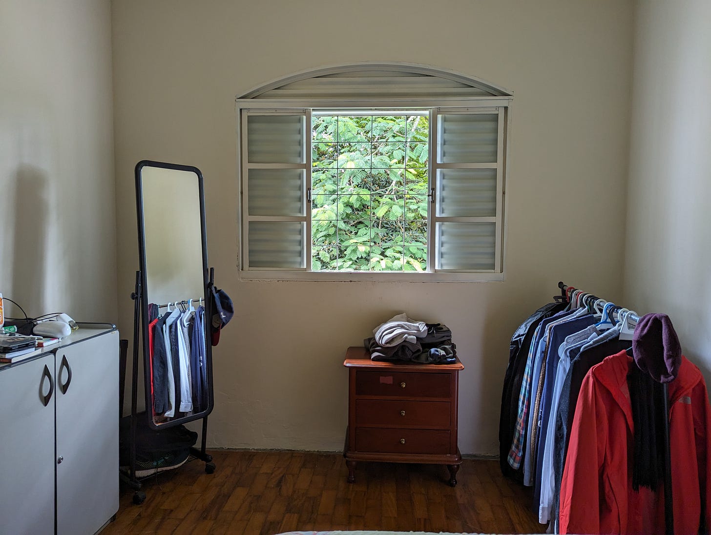 A bedroom, with a window at its centre showing green foliange from a tree. Beneath is is a wooden set of drawers with some clothes, to its right a rack with shirts and jackets, and to the left a mirror and a cupboard.