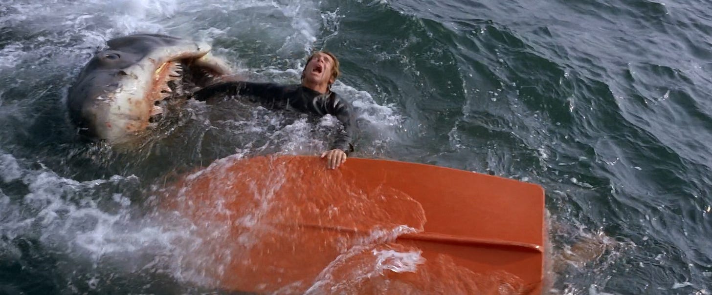 A frame from Jaws showing a terrified man clutching the top of a small, overturned rowboat with a red hull as he is dragged down by a gigantic shark.