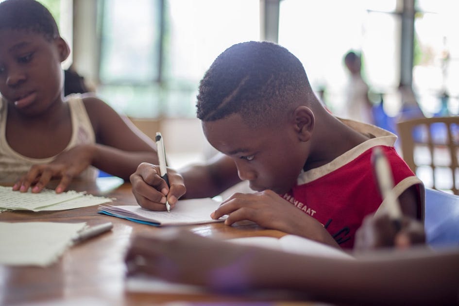 The image shows two children writing at a desk in their classroom