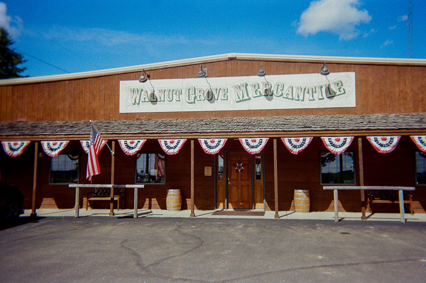 A “Walnut Grove Mercantile” old-timey store sign is displayed above a small patio roof lined with red, white, and blue banners. Two wooden hitching posts frame either side of the front door, creating a transition before reaching the tarred parking lot.
