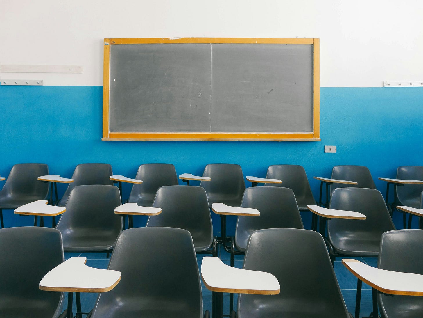 Photograph of an empty classroom. There are rows of desks and a black board.