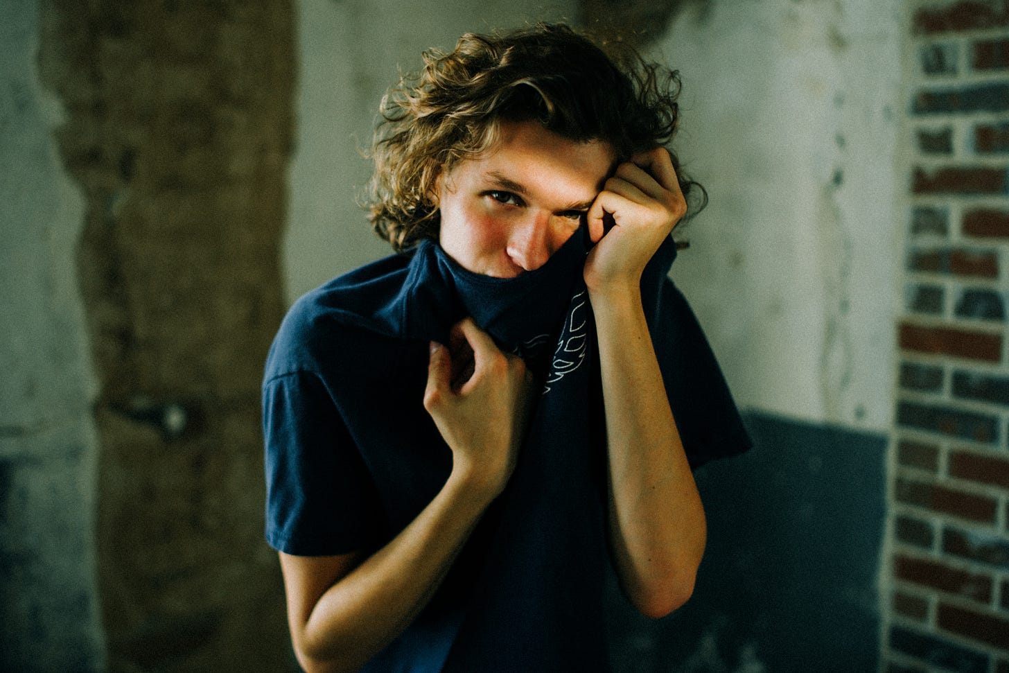a young white person, with blonde wavy hair, is wearing a blue shirt that they are pulling up around their face, as though trying to hide. They are standing in a run down building.