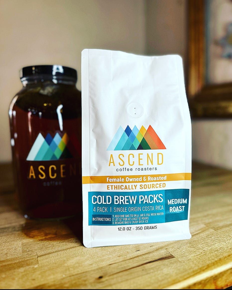 A white coffee bag labeled "Cold Brew Packs" sits on a wood countertop. A brown jar with a black lid is in the background.