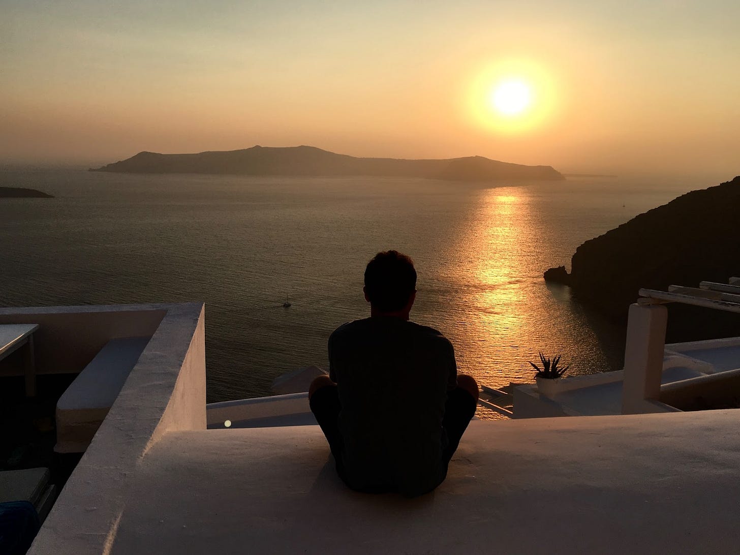 Un uomo di spalle, seduto sul gradino di un patio di una casa sul mare, osserva isole in lontananza al tramonto